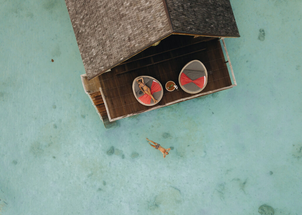 aerial blonde woman relaxing on day bed at sun terrace overwater suite man swimming looking up the sky in lagoon 1