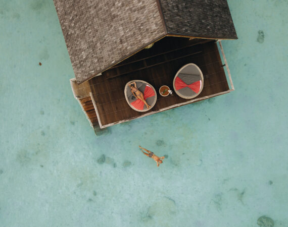 aerial blonde woman relaxing on day bed at sun terrace overwater suite man swimming looking up the sky in lagoon 1