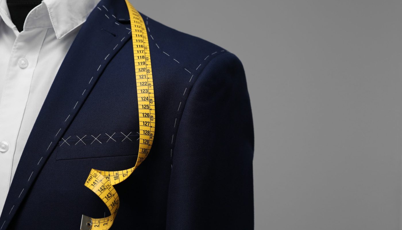 Semi-ready jacket with tailor's measuring tape on mannequin against grey background, closeup. Space for text